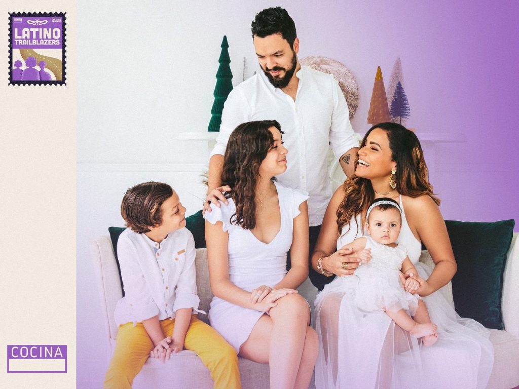 Cris Mercado, part of Latino Trailblazers, sitting a couch with her family, smiling. Her partner is standing behind the couch where she's stting along two children and a baby. Photo courtesy of Cris Mercado.