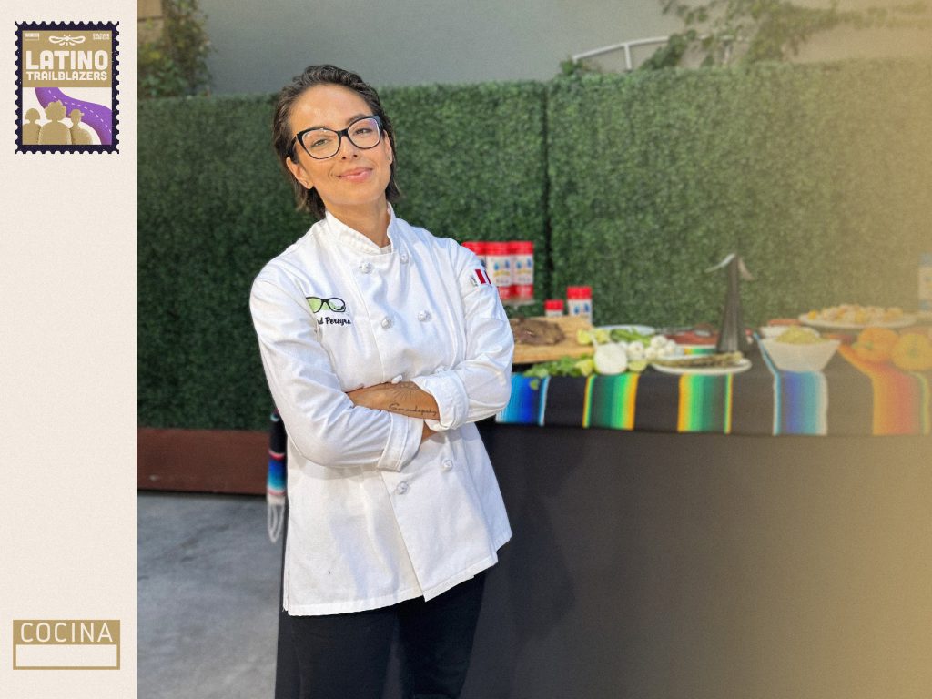 Chef Ingrid Pereyra, part of Latino Trailblazers, standing and smirking at the camera. She's wearing her chef apron, crossing her arms while posing in fron of a table filled with food.  Photo courtesy of Ingrid Pereyra.