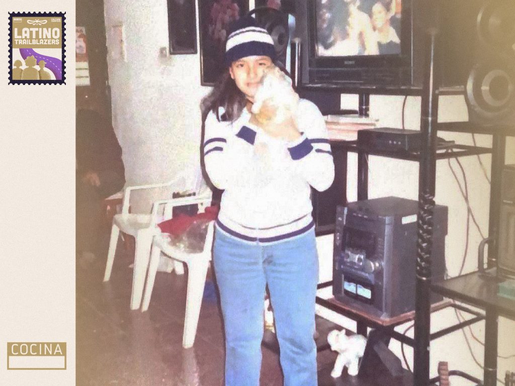 Chef Ingrid Pereyra, part of Latino, as a child. He's looking very seriously at the camera, holding a puppy in her arms, and wearing winter clothing. She's standing at a living room, with a TV set in the backdrop. Photo courtesy of Ingrid Pereyra.
